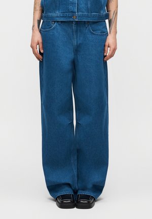 Personne portant un jean en denim bleu ample, des chaussures noires à bout carré, et un gilet en denim assorti, debout les mains détendues.