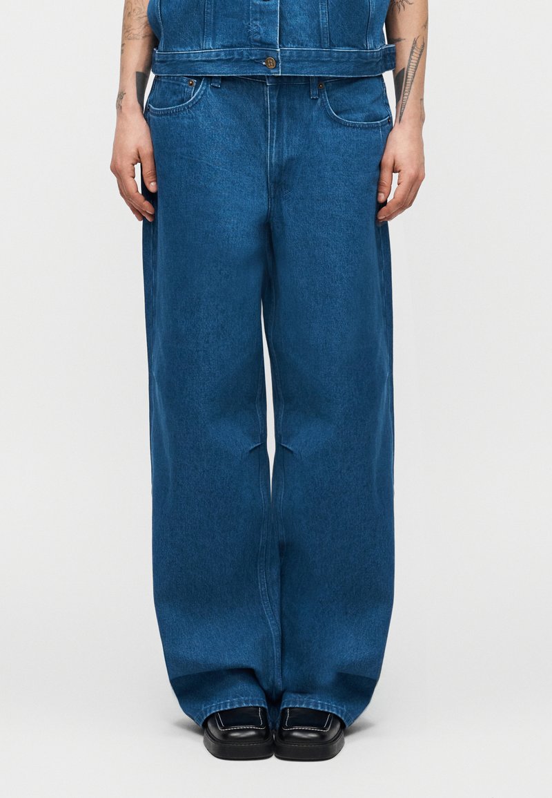 Άτομο που φοράει φαρδιά μπλε τζιν παντελόνια, μαύρα παπούτσια με τετράγωνη μύτη και τζιν γιλέκο που ταιριάζει, στέκεται με τα χέρια χαλαρά.