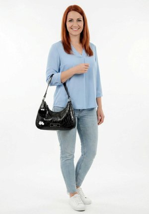 Vrouw met rood haar, gekleed in een lichtblauw blouse en spijkerbroek, houdt een zwarte handtas met decoratieve bedels vast en lacht naar de camera.