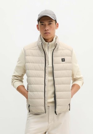Junger Mann mit Händen in den Taschen, trägt eine hellbeige gesteppte Weste, passenden Pullover, Hose und eine graue Kappe vor einfachem Hintergrund.