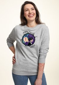 Donna che sorride in un felpa grigia con una grafica di due donne schiena contro schiena incorniciate da rami neri e ragnatele viola, indossando jeans blu.