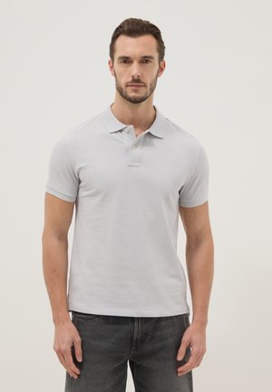 Uomo con capelli corti e scuri e barba, indossa una polo grigio chiaro e jeans grigio scuro, in piedi davanti a uno sfondo chiaro uniforme.