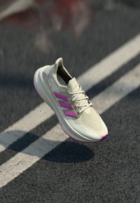 Beige löparskor med rosa accenter, mesh-överdel och en vadderad sula. Har tre distinkta sidosträck och en texturerad tåhuvud.