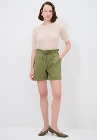 Maglione in maglia di colore beige chiaro con maniche corte; pantaloni shorts verde oliva a vita alta con cordoncino, orli piegati e tasche laterali; décolleté a punta.
