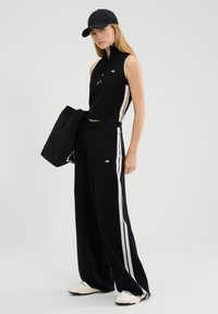 Schwarzes ärmelloses Oberteil mit Reißverschlusskragen, kombiniert mit hoch taillierter schwarzer Hose mit weißen Seitenstreifen. Trägt eine schwarze Mütze und hält eine Tasche.