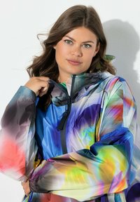 Chaqueta colorida de material ligero y brillante. Presenta un cuello alto, cierre de cremallera y patrones abstractos en tonos vibrantes de azul, rosa y verde.