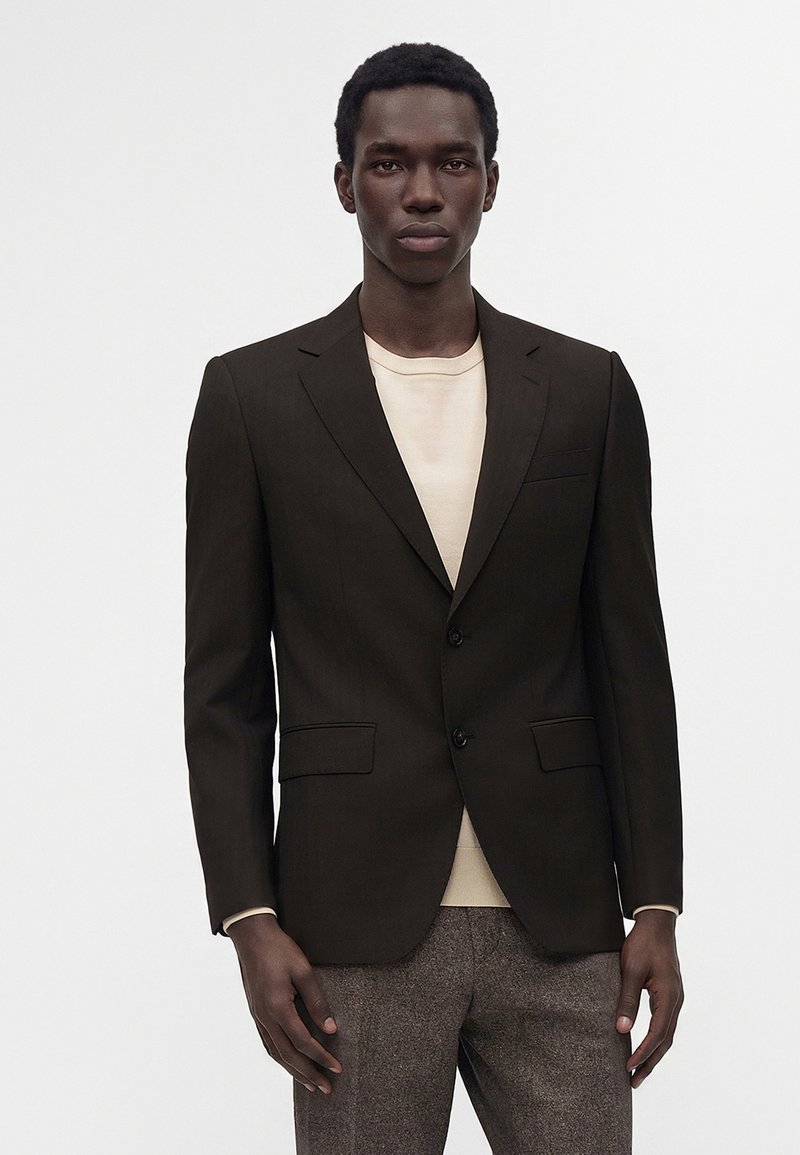 Homme portant un blazer noir, une chemise beige et un pantalon marron, debout devant un fond clair uni.