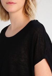 Černý, volný top s kulatým výstřihem a krátkými rukávy. Lehká, texturovaná tkanina s mírně průsvitným vzhledem.