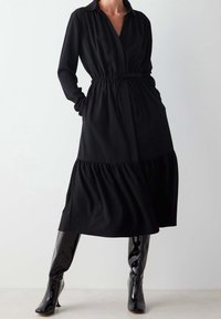 Nainen, joka käyttää mustaa pitkähihasta midi-mekkoa, jossa on kiristettävä vyötärö, ja kiiltävänmustia polvipituisia saappaita, seisoo yksinkertaista taustaa vasten.