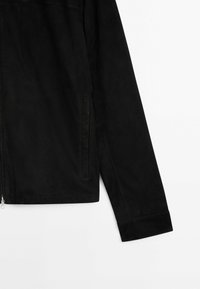 Détail de la manche et de la poche latérale d'une veste en daim noir sur fond blanc, montrant la fermeture éclair et les coutures.