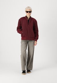 Burgundy bomber jacket with ribbed cuffs, zip closure, and slanted pockets. Paired with gray wide-leg jeans and black sneakers on a plain background.