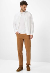 Camicia bianca button-up sopra una t-shirt bianca, abbinata a pantaloni beige e scarpe in suede marroni. I pantaloni hanno una vestibilità comoda con orli arrotolati.