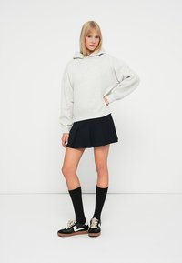 Felpa grigia chiaro con cappuccio, taglio oversize, texture morbida, abbinata a una gonna nera plissettata, calze nere al ginocchio e sneakers nere con accenti bianchi.