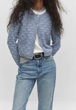 Personne portant un cardigan à boutons bleu clair texturé par-dessus un haut blanc avec ceinture et un jean bleu clair, mains dans le dos.