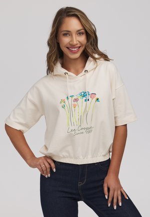 Uśmiechnięta kobieta nosi kremową krótką bluzę z kapturem z kwiatowym nadrukiem i napisem "Lee Cooper Since 1908", w połączeniu z ciemnoniebieskimi dżinsami.