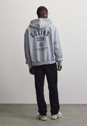 Mężczyzna stoi tyłem, ubrany w szarą bluzę z kapturem "Boxing Club", czarne spodnie, białe sneakersy i słuchawki, trzyma klucze przy boku.