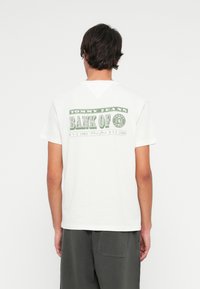 Fehér Tommy Jeans pólót viselő férfi, zöld "BANK OF" felirattal és sötét sorttal, háttal áll egy sima fehér háttér előtt.