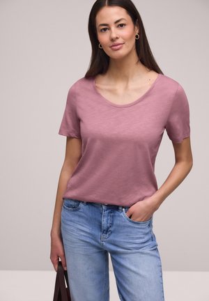 Ung kvinde iført en mauve kortærmet skjorte og lyseblå jeans, der holder en mørkebrun taske med den ene hånd i lommen.