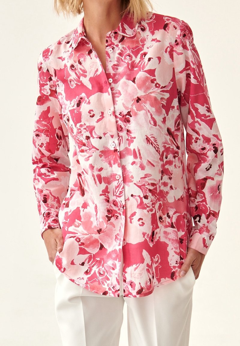 Femme portant une chemise à manches longues à boutons avec un motif floral rouge et rose, associée à un pantalon blanc, les mains dans les poches.