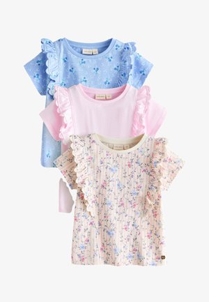 Jeune fille aux longs cheveux foncés portant un haut à volants à motif floral clair et un short assorti, des chaussettes blanches et des baskets crème, debout contre un mur uni.