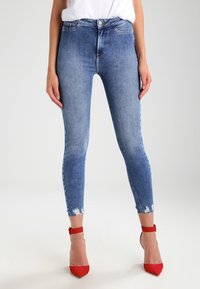 Höga blå denim skinny jeans med en urblekt finish, utrustade med råa fållar och en smal passform. Kombinerat med röda högklackade skor.