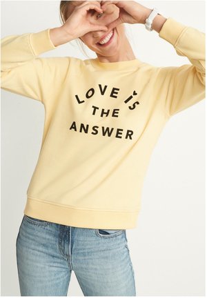 Lachend persoon met een geel sweatshirt met de tekst "LOVE IS THE ANSWER", maakt een hartvorm met de handen vlak bij het gezicht.