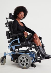 Γυναίκα με κοτσίδα αφέλειες, ντυμένη με μαύρο φόρεμα και μπότες, καθισμένη σε ένα μπλε ηλεκτρικό αναπηρικό αμαξίδιο, μπροστά σε λευκό απλό φόντο.