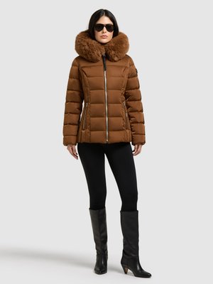 Le modèle porte une doudoune matelassée marron à col en fourrure, des lunettes de soleil noires, un legging et des bottes hautes jusqu’aux genoux, sur un fond uni.
