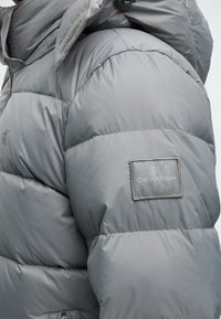 Doudoune gris clair avec capuche, coutures matelassées et patch logo Calvin Klein sur le haut du bras.
