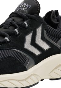 Zapatillas deportivas en negro y gris con un upper de tela texturizada, acentos de ante, un logo prominente y una suela blanca gruesa con ranuras.