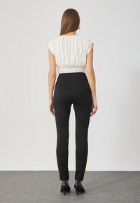 Λευκή, πλισέ μπλούζα με κοντά μανίκια σε συνδυασμό με μαύρο στενόμενο παντελόνι. Μαύρες ψηλοτάκουνες γόβες ολοκληρώνουν την εμφάνιση. Ουδέτερο φόντο.