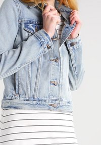 Ljusblå denimjacka med klassisk design, knäppning fram och två bröstfickor, bärs över en randig vit klänning.