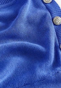 Blått stickat tyg med en slät textur, prytt med metallknappar dekorerade med klara rhinestones längs en kant.