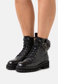 Botas de tobillo de cuero negro con una gruesa suela negra, con un diseño de cordones, herrajes en tono dorado y un acento decorativo de flor en el costado.