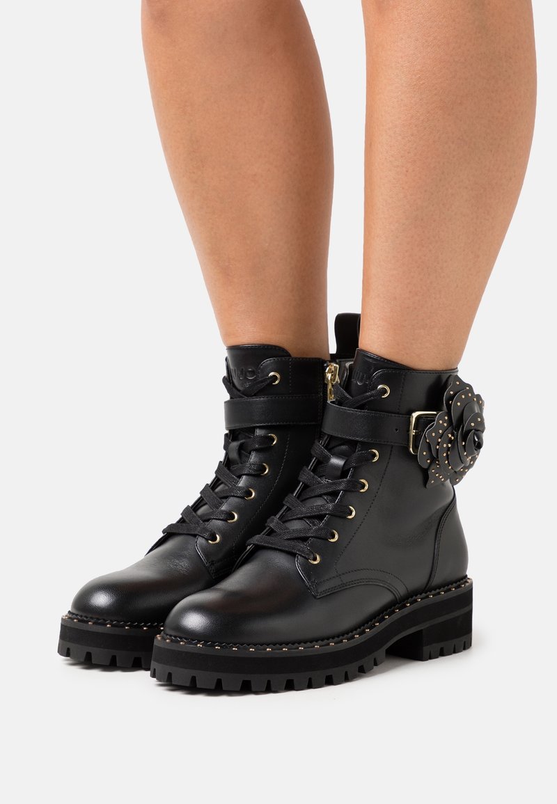 Botas de tobillo de cuero negro con una gruesa suela negra, con un diseño de cordones, herrajes en tono dorado y un acento decorativo de flor en el costado.