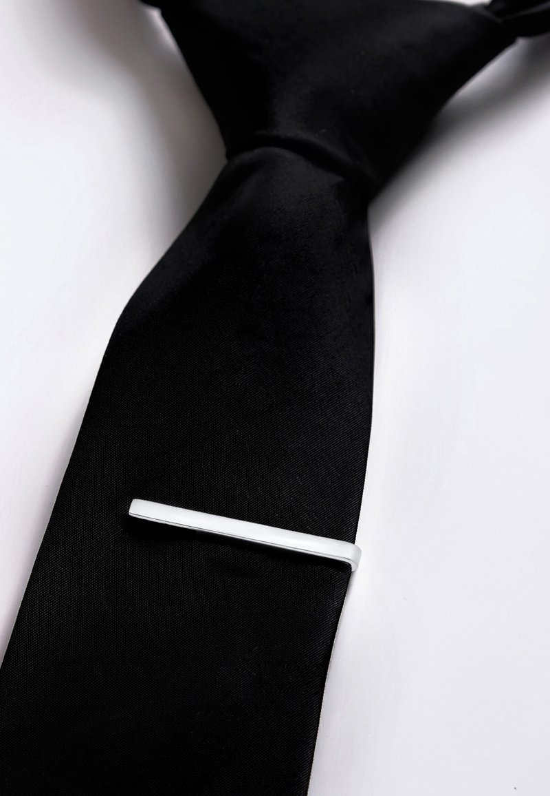 Cravatta in seta nera con una texture liscia, dotata di un fermacravatta in argento, di forma rettangolare, posizionato in diagonale sulla cravatta.