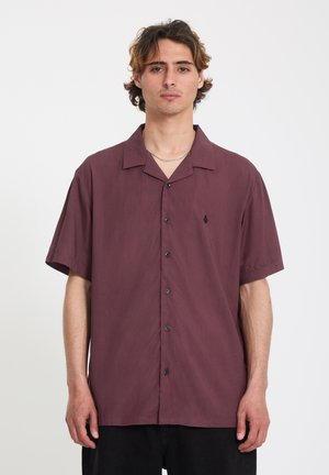 Ung mand med mellemlangt hår iført en kortærmet, bordeauxfarvet skjorte med knapper mod en ensfarvet baggrund.