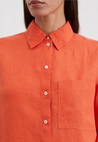 Orange linne skjorta med knappar, krage, framficka och vita knappar med subtil textur. Helt framifrån, inga ytterligare detaljer.