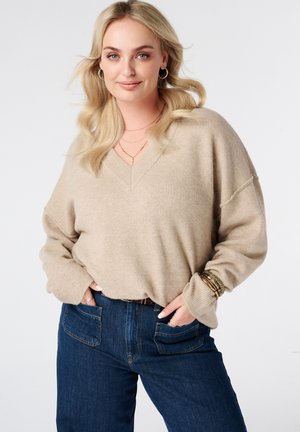 Blonde vrouw met losse golven draagt een beige V-hals trui en donkerblauwe jeans, aangevuld met gouden armbanden, ringen en creolen.