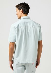 Lichtblauwe korte mouwen shirt met subtiele verticale strepen, een puntige kraag en een recht achterontwerp. Bevat een klein logo-label.