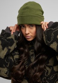 Zelený fleecový čiapky so zloženým lemom, skombinované s čiernou a olivovo camo fleecovou bundou. Textúrovaná tkanina a výrazné detaily vzoru.