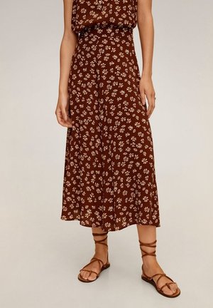 Kvinde iført en brun blomster-print midi-nederdel og matchende top kombineret med brune flade sandaler med remme på lys baggrund.