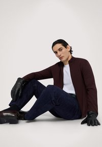 Burgundská zipová bunda, bílý tričko, tmavě modré semišové kalhoty, černé kožené rukavice a hnědé boty. Sedící postoj proti neutrálnímu pozadí.