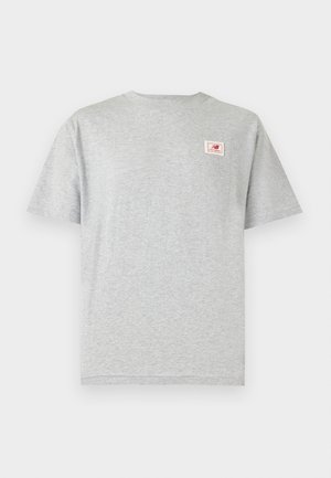 Mężczyzna z zaplecionymi włosami, ubrany w szary t-shirt New Balance i oliwkowe krótkie spodnie typu cargo, stoi tyłem na jednolitym białym tle.