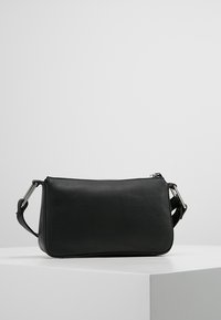 Small black leather handbag with adjustable shoulder strap and silver buckle, resting on a white pedestal against a plain background.