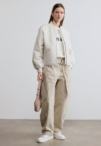 Ανοιχτό μπουφάν bomber με ραφές σε κρεμ απόχρωση, συνδυασμένο με λευκό πουλόβερ με μαύρο κείμενο, μπεζ παντελόνι με φαρδιά πόδια και λευκά αθλητικά παπούτσια. Συμπληρώνεται με τσάντα σε ταμπά απόχρωση.