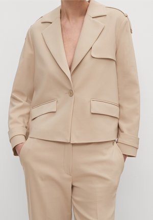 Veste beige cintrée avec larges revers, poches à rabat, fermeture par un bouton, portée avec un pantalon assorti et les mains dans les poches.