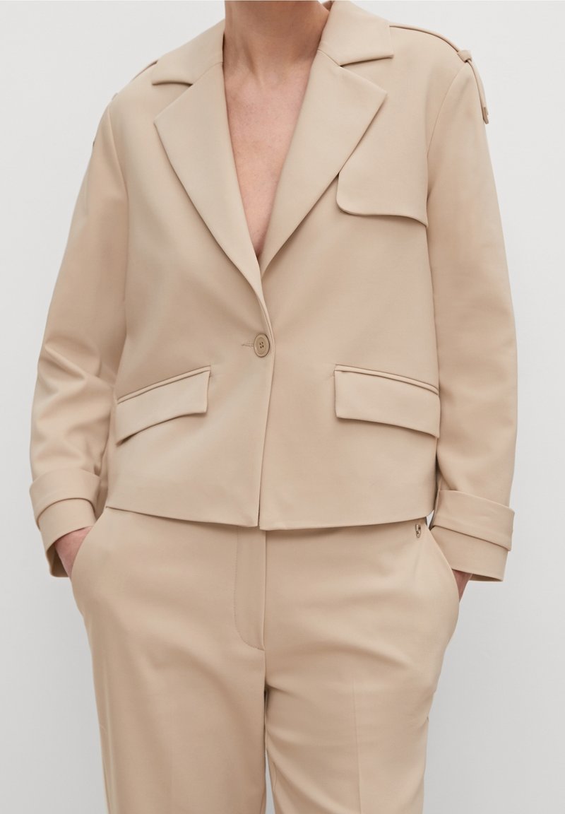 Veste beige cintrée avec larges revers, poches à rabat, fermeture par un bouton, portée avec un pantalon assorti et les mains dans les poches.