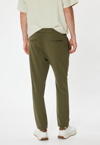 Olivgrüne, schmal zulaufende Hose mit Kordelzugbund, zwei Gesäßtaschen und feiner Textur. Kombiniert mit weißen Schuhen mit braunen Akzenten.