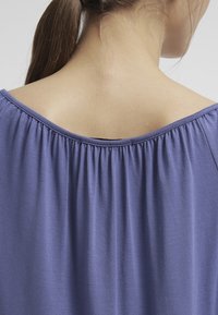 Haut en tissu élastique violet avec un large col et un dos froncé, montrant une texture douce et une finition lisse.